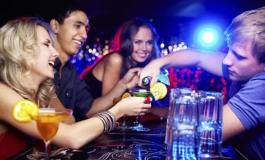 Four young adults at a lively bar in Punta Cana, smiling and laughing as a bartender pours drinks. Colorful cocktails with fruit garnishes sit on the table, and hanging lights capture the festive energy of Punta Cana nightlife.