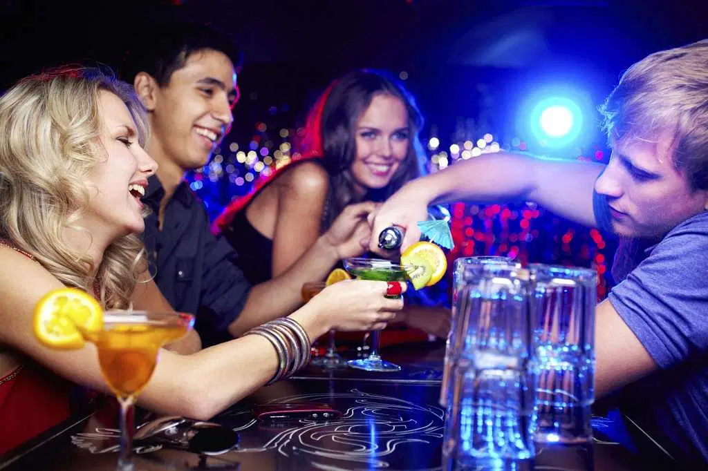 Four young adults at a lively bar in Punta Cana, smiling and laughing as a bartender pours drinks. Colorful cocktails with fruit garnishes sit on the table, and hanging lights capture the festive energy of Punta Cana nightlife.