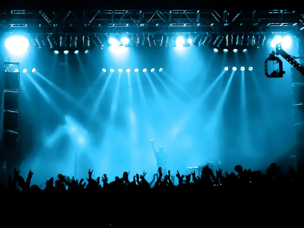 A crowd at a concert cheers with raised hands before a brightly lit stage, embodying the vibrant Punta Cana nightlife. Blue spotlights beam above as a performer stands center stage, silhouetted against the lights and holding a microphone.