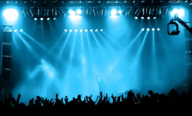 A crowd at a concert cheers with raised hands before a brightly lit stage, embodying the vibrant Punta Cana nightlife. Blue spotlights beam above as a performer stands center stage, silhouetted against the lights and holding a microphone.