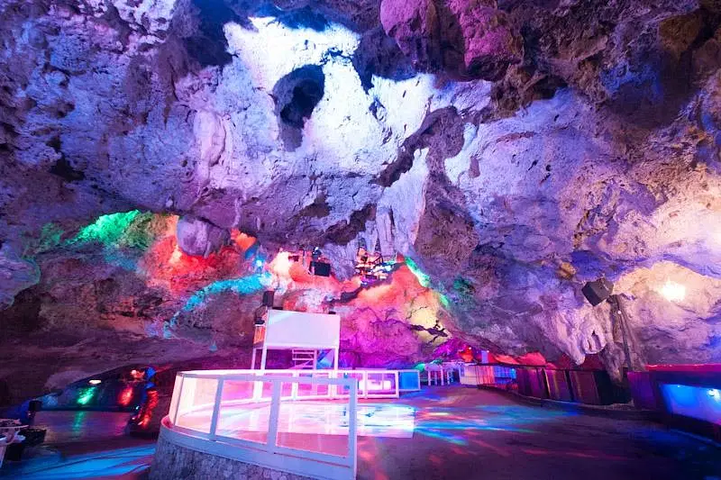 A vibrant nightclub set inside a natural cave, Imagine Punta Cana boasts colorful lights illuminating rocky ceilings and walls. A white DJ booth, reflective floors, and unique seating areas showcase the best of Punta Cana nightlife.