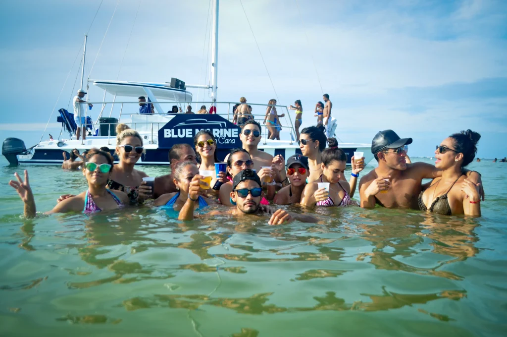 A group of people in swimsuits stand in shallow ocean water, holding drinks and smiling during one of the best tours in Punta Cana. Behind them, the "BLUE MARINE" boat floats under a clear sky, with more guests enjoying their sunny Punta Cana excursion.