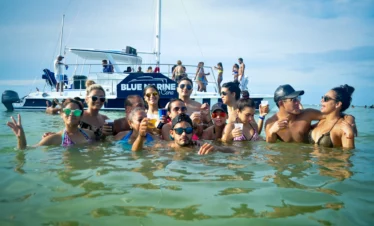 A group of people in swimsuits stand in shallow ocean water, holding drinks and smiling during one of the best tours in Punta Cana. Behind them, the "BLUE MARINE" boat floats under a clear sky, with more guests enjoying their sunny Punta Cana excursion.