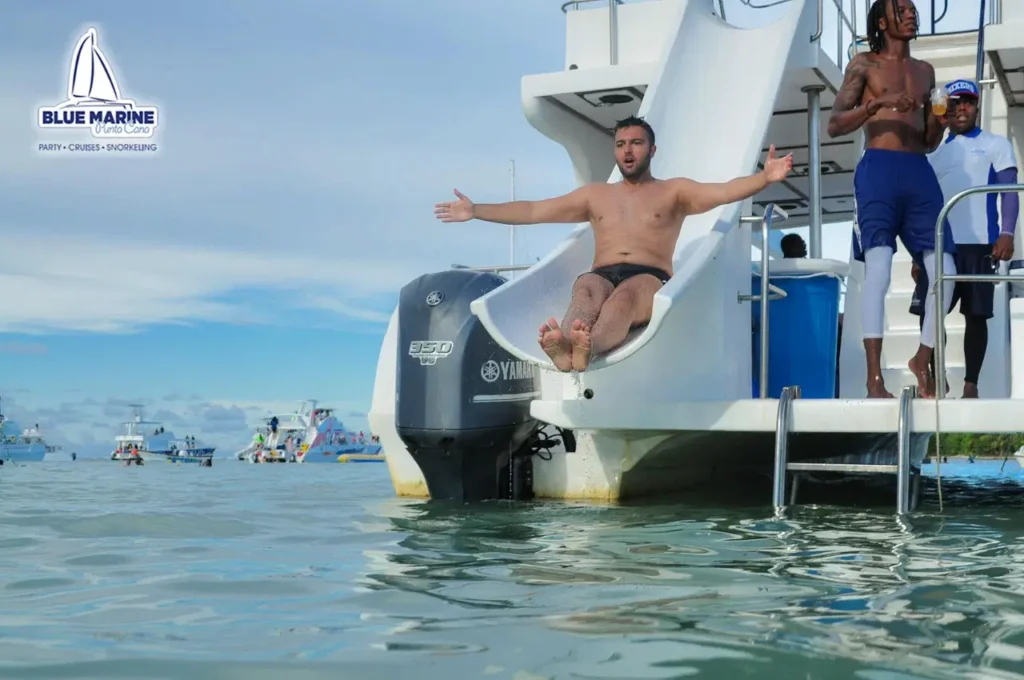 A shirtless man slides down a waterslide attached to a boat labeled "Blue Marine," arms open wide, about to splash into the sea. Enjoy exciting Punta Cana excursions like this, with other people and boats in the calm blue waters under a sunny sky.