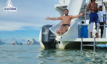 A shirtless man slides down a waterslide attached to a boat labeled "Blue Marine," arms open wide, about to splash into the sea. Enjoy exciting Punta Cana excursions like this, with other people and boats in the calm blue waters under a sunny sky.