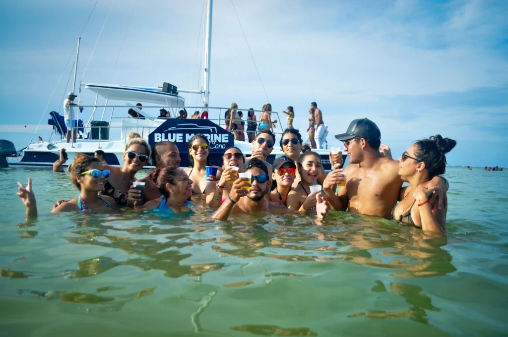 A group of people stand in shallow ocean water, laughing and holding drinks during one of the popular Punta Cana tours. Behind them, a white catamaran with the sign "BLUE MARINE" is anchored as more guests enjoy the sunny day on board.