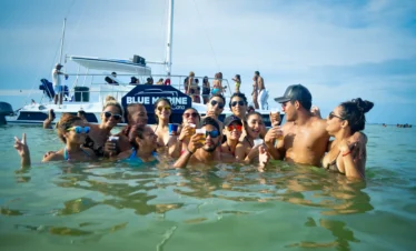 A group of people stand in shallow ocean water, laughing and holding drinks during one of the popular Punta Cana tours. Behind them, a white catamaran with the sign "BLUE MARINE" is anchored as more guests enjoy the sunny day on board.