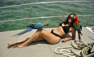 A woman in a black bikini relaxes on a boat deck near the ocean during one of her Punta Cana tours. Smiling, she enjoys a red-and-green parrot nuzzling her face and a blue-and-yellow parrot perched on her thigh, with coiled ropes and metal railing nearby.