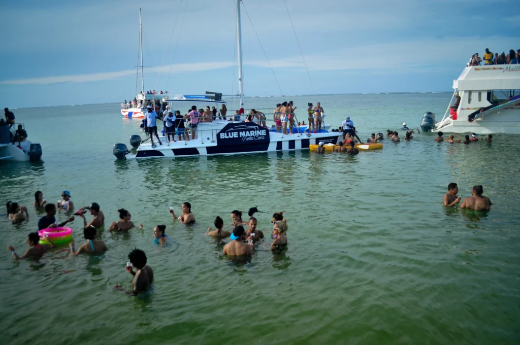 A group enjoys festive tours in Punta Cana, swimming and relaxing in shallow green water near two "BLUE MARINE" boats. Others lounge on deck or join the fun in the water, all under a cloudy sky. The lively scene captures the spirit of Punta Cana tours.