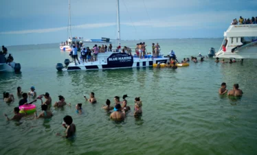 A group enjoys festive tours in Punta Cana, swimming and relaxing in shallow green water near two "BLUE MARINE" boats. Others lounge on deck or join the fun in the water, all under a cloudy sky. The lively scene captures the spirit of Punta Cana tours.