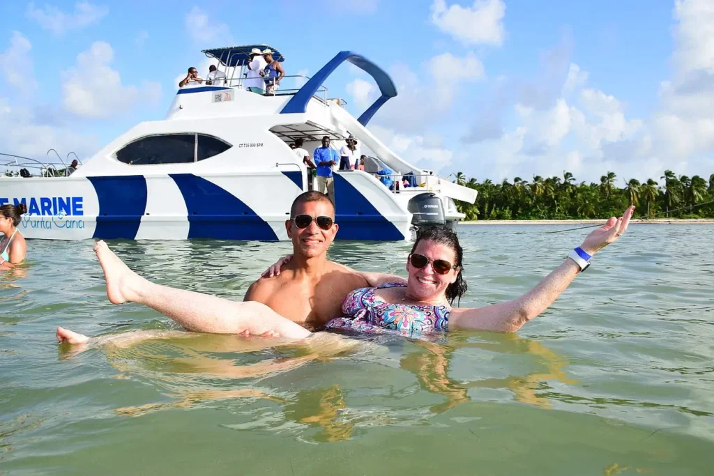 A smiling man and woman relax in shallow, clear water during their Punta Cana trip, with a large white and blue catamaran anchored behind them. Palm trees and a blue sky complete this idyllic scene.
