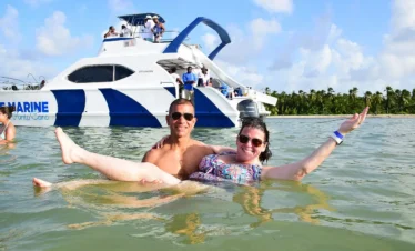 A smiling man and woman relax in shallow, clear water during their Punta Cana trip, with a large white and blue catamaran anchored behind them. Palm trees and a blue sky complete this idyllic scene.