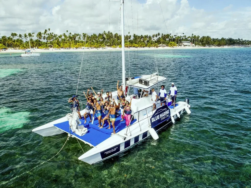 A group of people in swimwear pose cheerfully on the “Blue Marine” catamaran, anchored over turquoise water. Palm trees and a sandy beach set the scene—perfect for unforgettable Punta Cana excursions or your next Punta Cana trip.