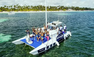 A group of people in swimwear pose cheerfully on the “Blue Marine” catamaran, anchored over turquoise water. Palm trees and a sandy beach set the scene—perfect for unforgettable Punta Cana excursions or your next Punta Cana trip.