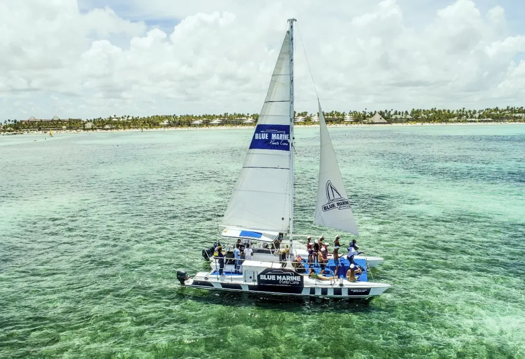 A catamaran labeled "Blue Marine" sails over clear, shallow turquoise water near a tropical shoreline lined with palm trees. Guests on board, enjoying the sunny day, experience one of the top Punta Cana excursions under partly cloudy skies.