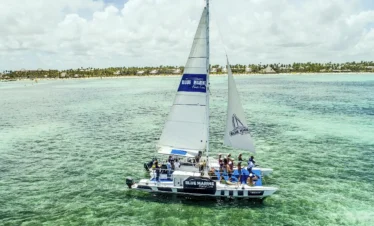 A catamaran labeled "Blue Marine" sails over clear, shallow turquoise water near a tropical shoreline lined with palm trees. Guests on board, enjoying the sunny day, experience one of the top Punta Cana excursions under partly cloudy skies.