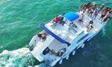 Aerial view of a white catamaran with a blue canopy cruising on clear turquoise water. Perfect for tours in Punta Cana, several people enjoy the sun and sea on the front deck as the boat’s wake trails behind.