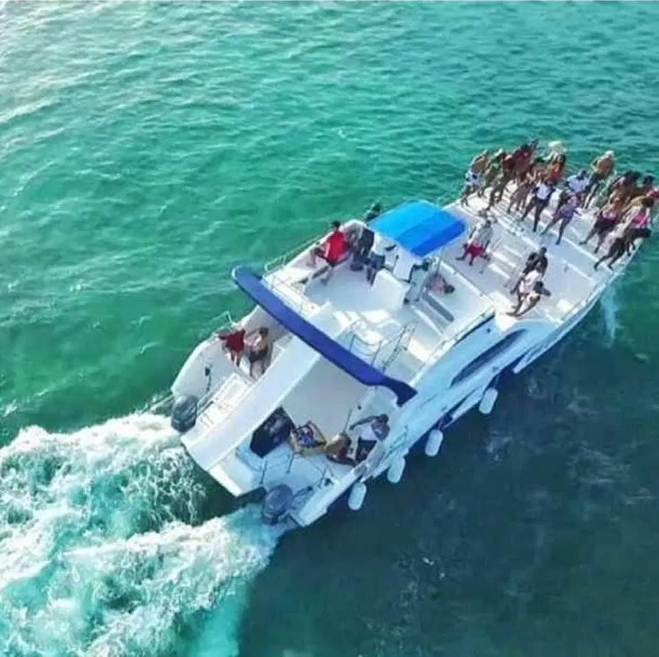 Aerial view of a white catamaran with a blue canopy cruising on clear turquoise water. Perfect for tours in Punta Cana, several people enjoy the sun and sea on the front deck as the boat’s wake trails behind.