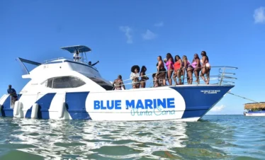 A group of women in colorful swimsuits stand together on the bow of a white and blue yacht labeled "BLUE MARINE Punta Cana," enjoying clear water and sunny skies—a perfect scene for unforgettable tours en Punta Cana. A crew member stands at the rear.