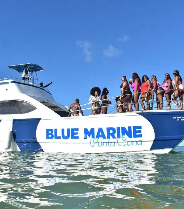 A group of women in colorful swimsuits stand together on the bow of a white and blue yacht labeled "BLUE MARINE Punta Cana," enjoying clear water and sunny skies—a perfect scene for unforgettable tours en Punta Cana. A crew member stands at the rear.