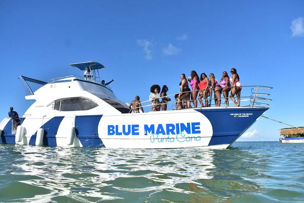 A group of women in colorful swimsuits stand together on the bow of a white and blue yacht labeled "BLUE MARINE Punta Cana," enjoying clear water and sunny skies—a perfect scene for unforgettable tours en Punta Cana. A crew member stands at the rear.