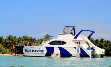 A white catamaran with “BLUE MARINE” on the side floats near a palm-lined shore. Several people enjoy swimming in the clear, turquoise water—an unforgettable scene from tours in Punta Cana under a bright blue sky.
