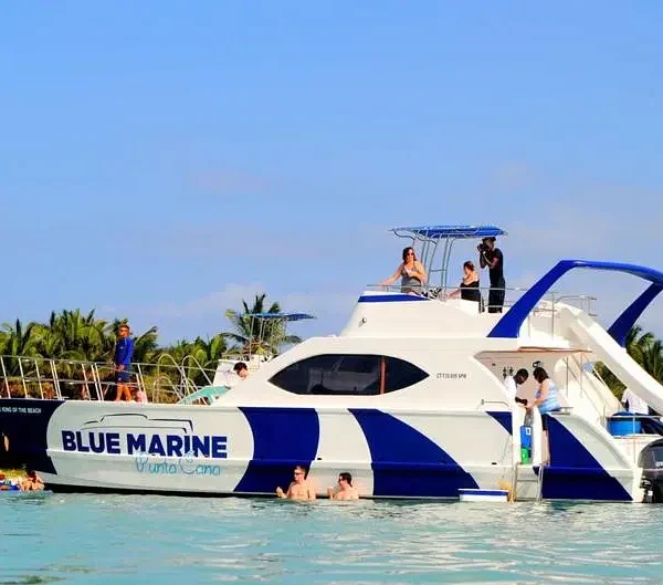 A white catamaran with “BLUE MARINE” on the side floats near a palm-lined shore. Several people enjoy swimming in the clear, turquoise water—an unforgettable scene from tours in Punta Cana under a bright blue sky.