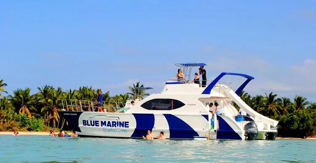 A white catamaran with “BLUE MARINE” on the side floats near a palm-lined shore. Several people enjoy swimming in the clear, turquoise water—an unforgettable scene from tours in Punta Cana under a bright blue sky.