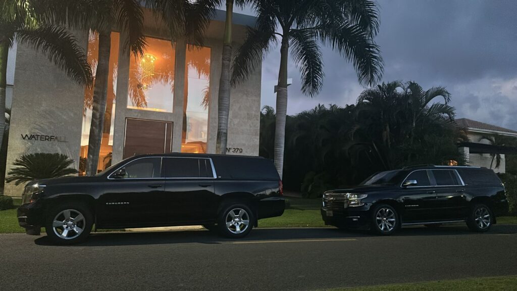 Two large black SUVs are parked side by side at dusk in front of a modern house with tall windows and lush greenery—reminiscent of exclusive tours in Punta Cana. The warmly lit "WATERFALL" sign enhances the tropical, inviting vibe.