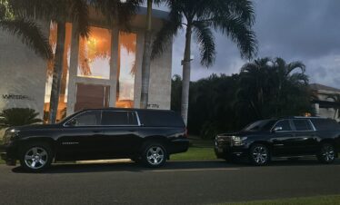 Two large black SUVs are parked side by side at dusk in front of a modern house with tall windows and lush greenery—reminiscent of exclusive tours in Punta Cana. The warmly lit "WATERFALL" sign enhances the tropical, inviting vibe.