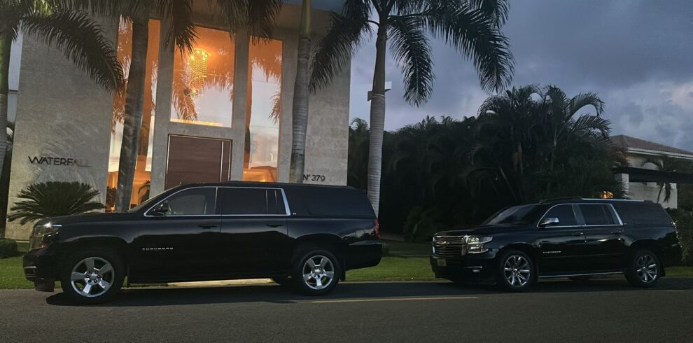 Two large black SUVs are parked side by side at dusk in front of a modern house with tall windows and lush greenery—reminiscent of exclusive tours in Punta Cana. The warmly lit "WATERFALL" sign enhances the tropical, inviting vibe.