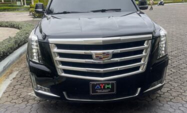 A black Cadillac Escalade SUV is parked on a cobblestone driveway surrounded by palm trees and tropical greenery under a cloudy sky. Perfect for a VIP transfer in Punta Cana, the SUV faces the camera with a decorative “ATM” license plate visible.