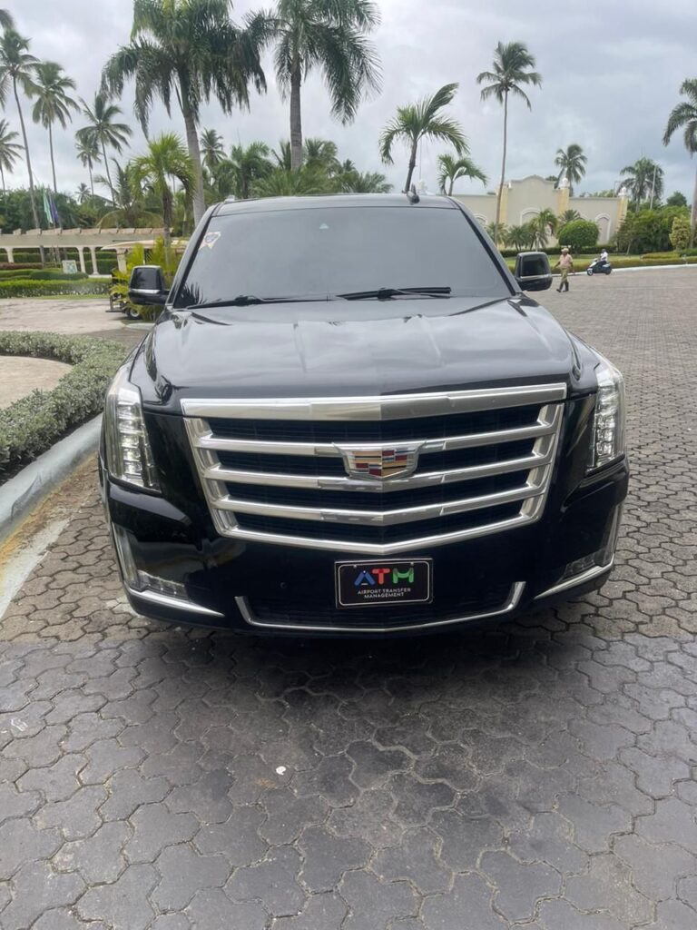A black Cadillac Escalade SUV is parked on a cobblestone driveway surrounded by palm trees and tropical greenery under a cloudy sky. Perfect for a VIP transfer in Punta Cana, the SUV faces the camera with a decorative “ATM” license plate visible.