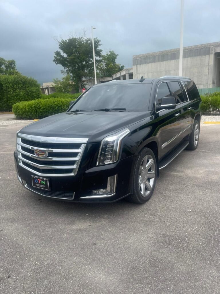 A black Cadillac Escalade SUV, perfect for VIP transfer, is parked outdoors with tinted windows, chrome accents, large rims, and a bold Cadillac emblem. Trees, a concrete building, and a cloudy sky frame the Punta Cana setting.