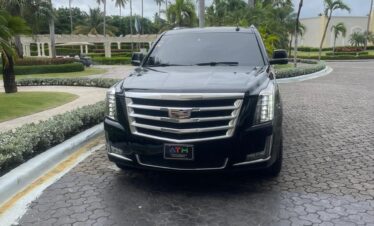 A black Cadillac Escalade SUV, ideal for a VIP transfer in Punta Cana, is parked on a circular cobblestone driveway, surrounded by palm trees and manicured bushes. The tinted vehicle faces the camera with an elegant building under a cloudy sky in the background.