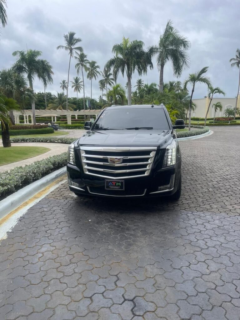 A black Cadillac Escalade SUV, ideal for a VIP transfer in Punta Cana, is parked on a circular cobblestone driveway, surrounded by palm trees and manicured bushes. The tinted vehicle faces the camera with an elegant building under a cloudy sky in the background.