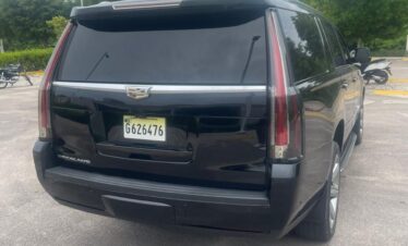 A black Cadillac Escalade SUV, often used for VIP transfer services in Punta Cana, is parked in an outdoor lot on a cloudy day. The vehicle features tinted windows and a visible rear license plate. Trees and motorcycles line the paved background.