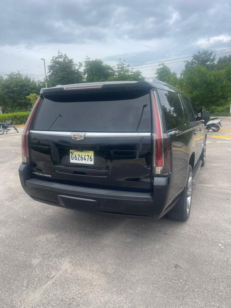 A black Cadillac Escalade SUV, often used for VIP transfer services in Punta Cana, is parked in an outdoor lot on a cloudy day. The vehicle features tinted windows and a visible rear license plate. Trees and motorcycles line the paved background.
