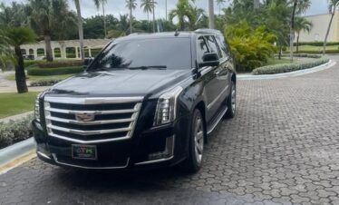 A shiny black Cadillac Escalade SUV, perfect for a VIP transfer, is parked on a circular brick driveway lined with palm trees and lush greenery. The sky is cloudy, and a grand Punta Cana building with arches and white columns stands behind.