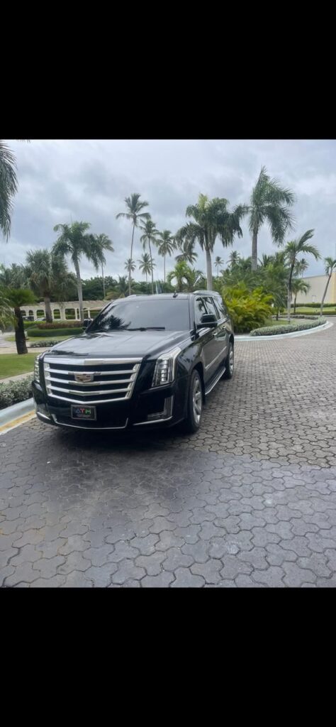 A shiny black Cadillac Escalade SUV, perfect for a VIP transfer, is parked on a circular brick driveway lined with palm trees and lush greenery. The sky is cloudy, and a grand Punta Cana building with arches and white columns stands behind.