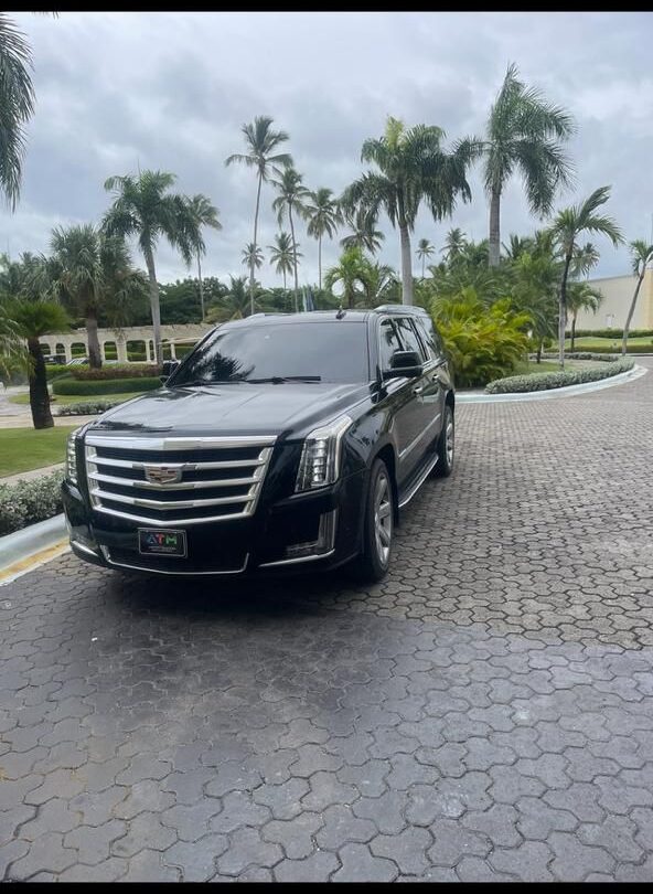 A shiny black Cadillac Escalade SUV, perfect for a VIP transfer, is parked on a circular brick driveway lined with palm trees and lush greenery. The sky is cloudy, and a grand Punta Cana building with arches and white columns stands behind.