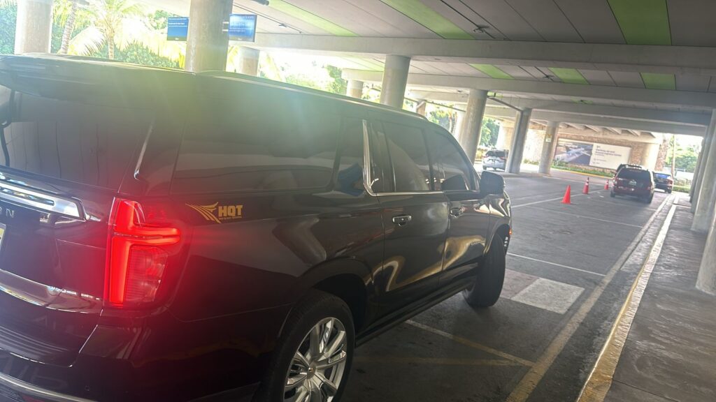 A black SUV with tinted windows and an “HQT” logo awaits a VIP transfer at Punta Cana’s covered airport pickup zone. Sunlight filters in, reflecting off the car, as orange cones, pillars, and a green-striped ceiling frame the scene.