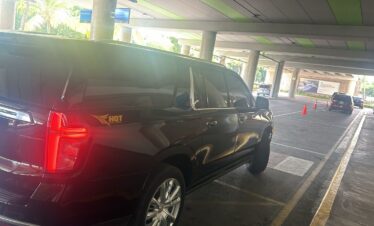 A black SUV with tinted windows and an “HQT” logo awaits a VIP transfer at Punta Cana’s covered airport pickup zone. Sunlight filters in, reflecting off the car, as orange cones, pillars, and a green-striped ceiling frame the scene.