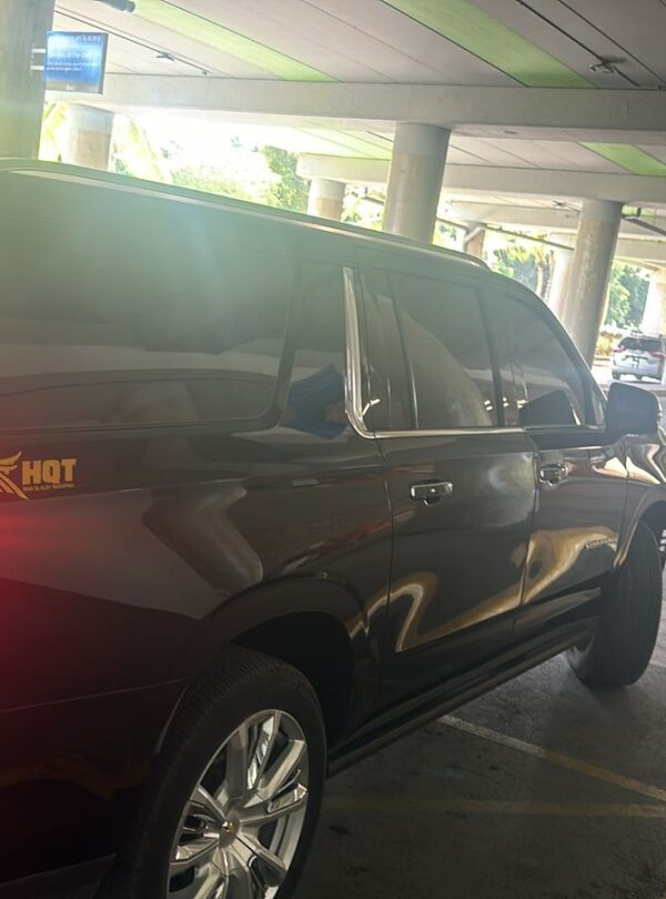A black SUV with tinted windows and an “HQT” logo awaits a VIP transfer at Punta Cana’s covered airport pickup zone. Sunlight filters in, reflecting off the car, as orange cones, pillars, and a green-striped ceiling frame the scene.