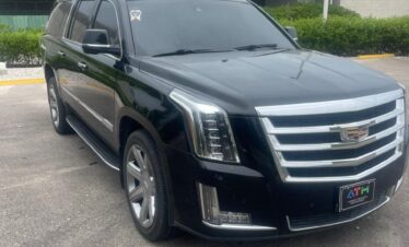 A black Cadillac Escalade SUV, ideal for VIP transfer in Punta Cana, is parked on a paved road near a low building with glass windows and green shrubs. The sky is overcast, highlighting the vehicle’s tinted windows, chrome trim, and bold Cadillac grille.