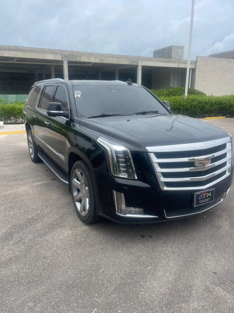 A black Cadillac Escalade SUV, ideal for VIP transfer in Punta Cana, is parked on a paved road near a low building with glass windows and green shrubs. The sky is overcast, highlighting the vehicle’s tinted windows, chrome trim, and bold Cadillac grille.