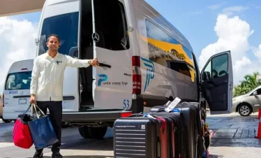 A smiling man stands beside an open van door labeled “Punta Cana Trip,” holding two bags. Suitcases line the pavement, ready for exciting tours in Punta Cana under the sunny blue sky, with another vehicle in the background.