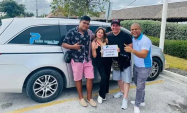 Four people stand smiling by a silver van in a parking lot, holding a “Bienvenido Patron Javier” sign. The happy group is ready for their Punta Cana excursions, casually dressed with lush greenery and buildings in the background.