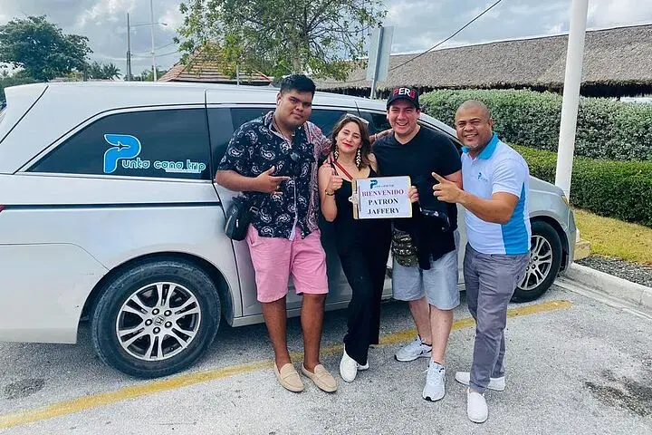 Four people stand smiling by a silver van in a parking lot, holding a “Bienvenido Patron Javier” sign. The happy group is ready for their Punta Cana excursions, casually dressed with lush greenery and buildings in the background.