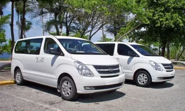Two white Hyundai vans, perfect for tours in Punta Cana, are parked side by side in a sunny outdoor lot. Green trees, blue water, and a clear sky set the scene. Both vans have tinted windows and face right, ready for your next Punta Cana trip.